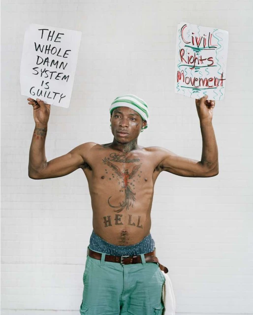 Shirtless man holds protest signs with text against a white brick background, wearing a cap and casual shorts with visible tattoos. 