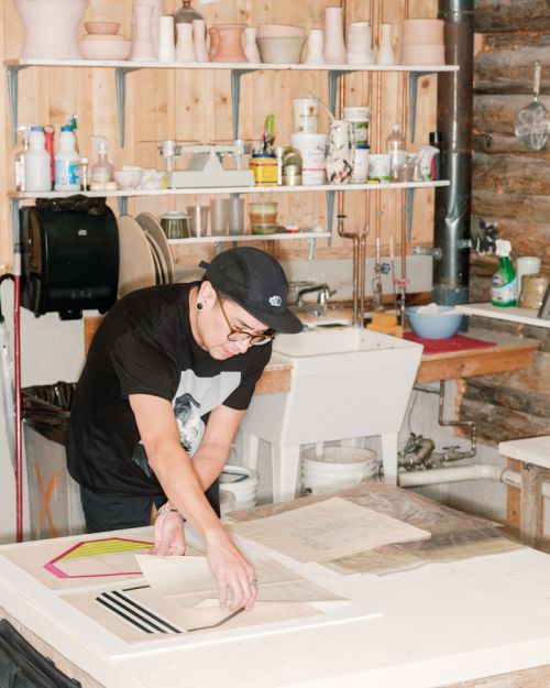 Person in a workshop with art supplies and ceramic objects on shelves, working with geometric designs and drafting on paper.