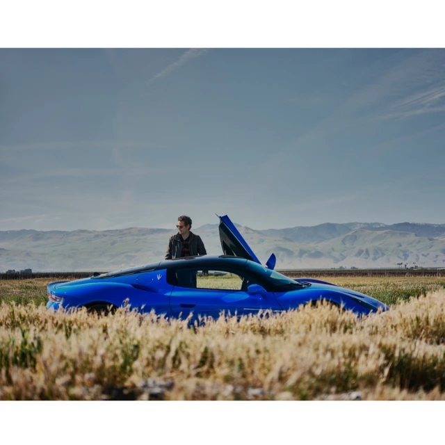 Blue Maserati sports car parked in a field with an adult male standing alongside it with mountains and blue skies in the background.