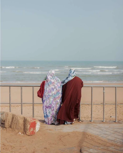 Two adult females in traditional dress stand on a sandy beach by a railing, overlooking the ocean with small waves under a bright sky.