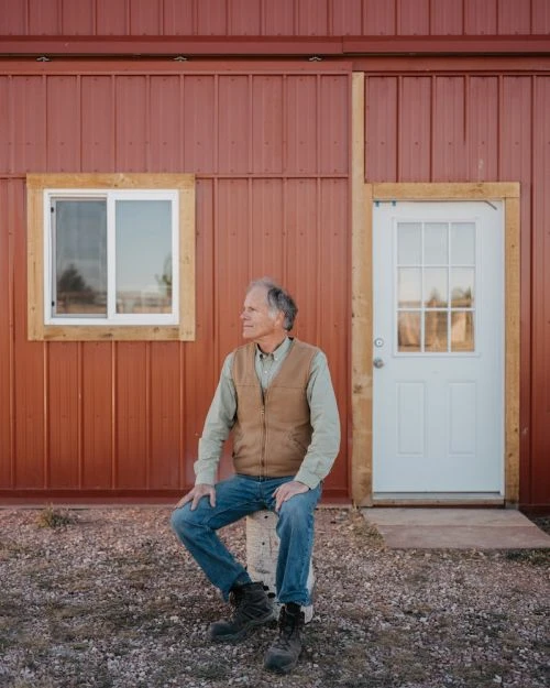 An adult male sits on a concrete block outside a red metal building on a gravel surface, dressed in a vest, shirt, jeans, and boots.