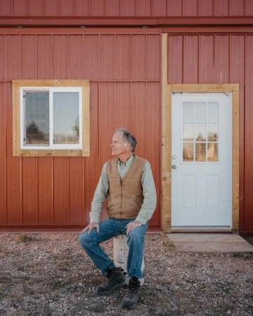 An adult male sits on a concrete block outside a red metal building on a gravel surface, dressed in a vest, shirt, jeans, and boots.