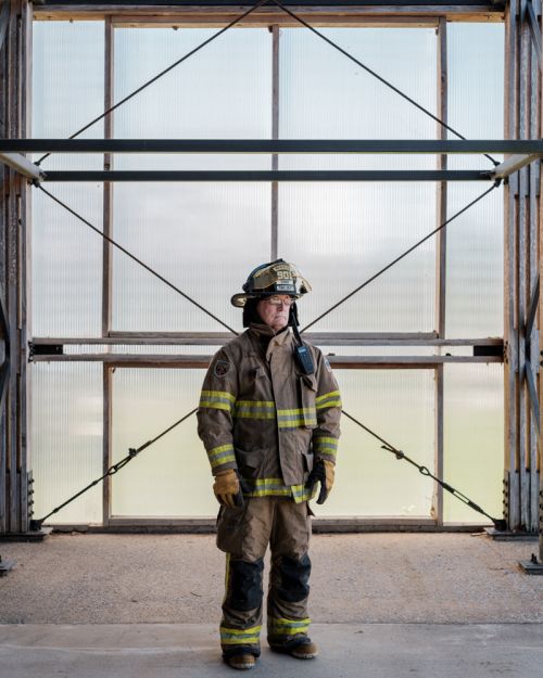 Full shot of adult firefighter standing in gear against a translucent window and metal frame structure in natural outdoor light.