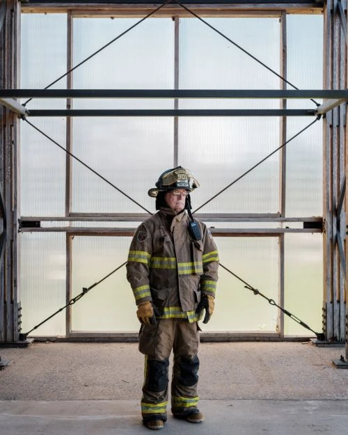 Full shot of adult firefighter standing in gear against a translucent window and metal frame structure in natural outdoor light.