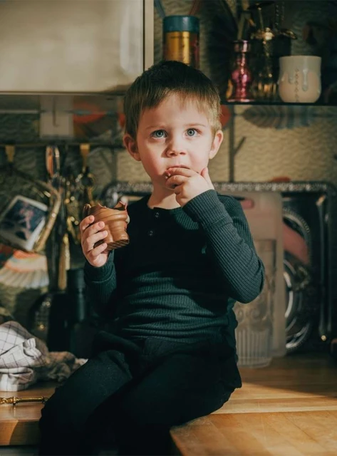 Young boy in dark clothing sits on wooden surface, eating treat from pot-shaped container in a brightly lit indoor environment.