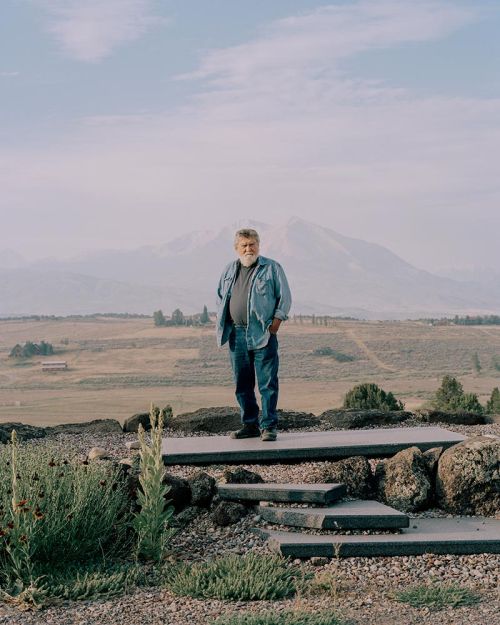Full-length portrait of an older man standing on concrete steps outdoors, wearing jeans and a blue denim shirt, against a mountain backdrop.