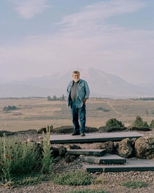 Full-length portrait of an older man standing on concrete steps outdoors, wearing jeans and a blue denim shirt, against a mountain backdrop.