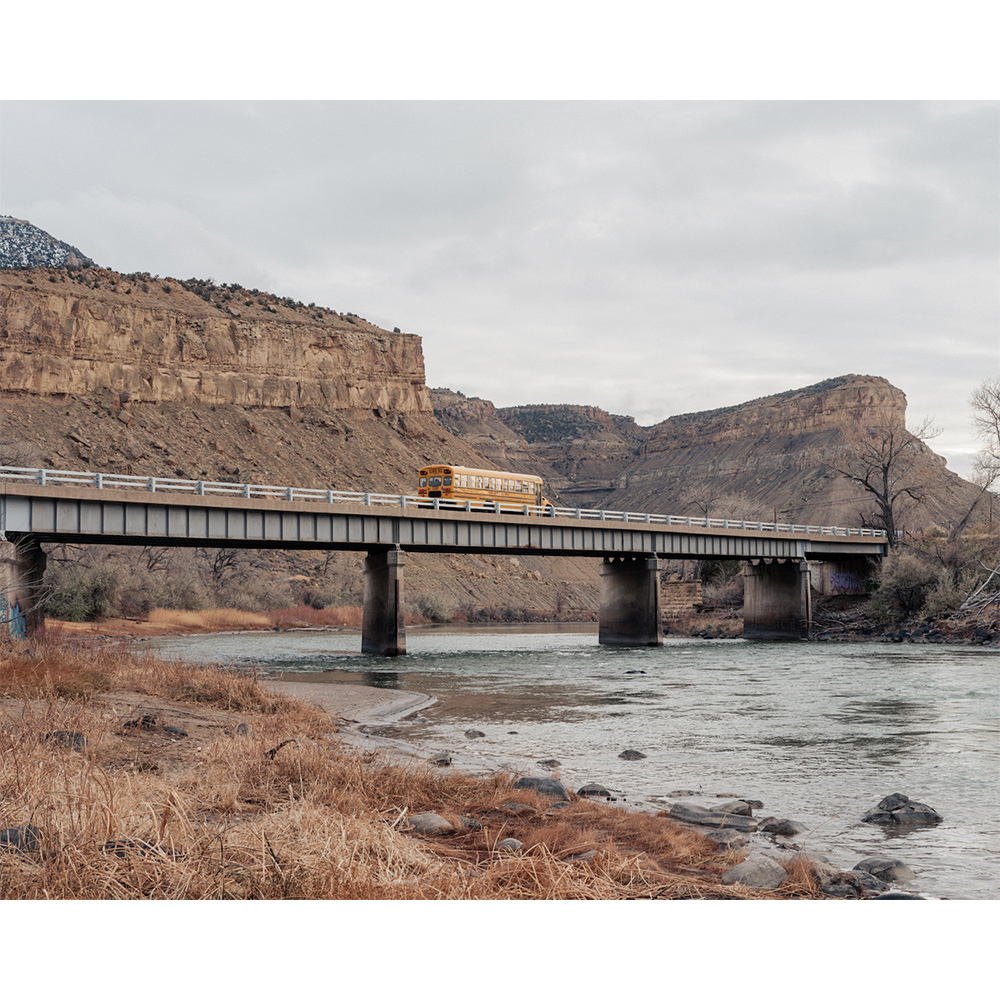 A yellow school bus crosses a steel bridge over a river, with canyons and a cloudy sky in the background, creating a scenic composition.