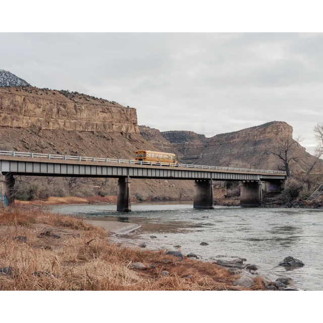 A yellow school bus crosses a steel bridge over a river, with canyons and a cloudy sky in the background, creating a scenic composition.
