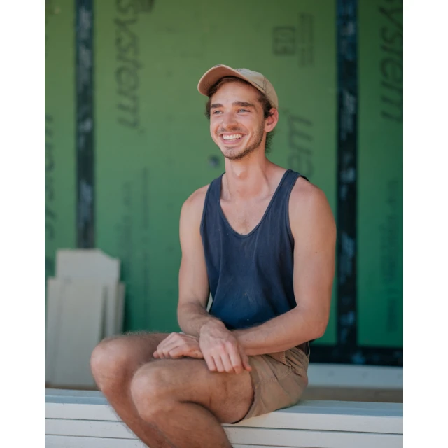 Smiling man in a tank top and hat sits outside. He has brown hair and a goatee, and the background features green building material.