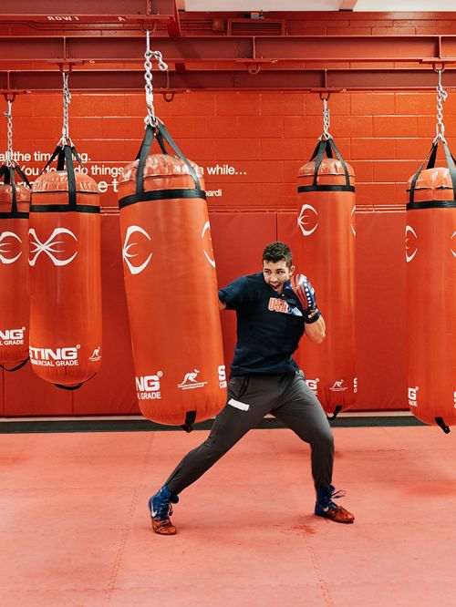 Man in boxing stance by a punching bag in a gym with red walls. He is wearing boxing gloves and athletic wear.