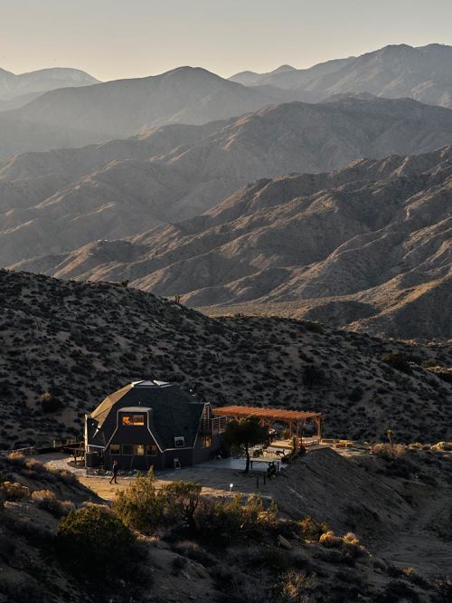 Exterior view of a modern home nestled in a mountainous desert landscape, illuminated by soft sunlight, showcasing architecture and environment.