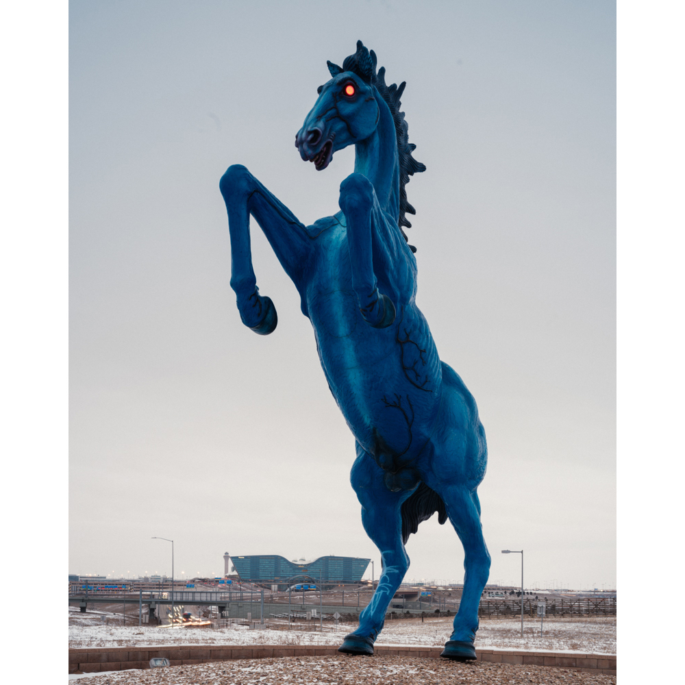 Monumental sculpture of a blue horse rearing on its hind legs, situated on a stone base with a building and clear sky in the background.