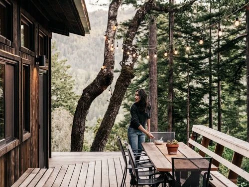 Woman on a wooden deck of a house near a forest with string lights, with an outdoor table, chairs, and potted plants visible.