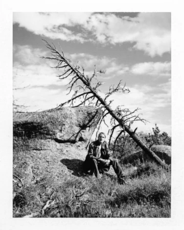Black and white portrait of a young adult sitting on a rock with a dead tree, set against a sky with dramatic clouds.