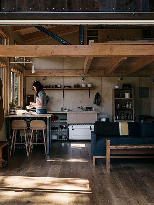 Woman stands in a modern cabin kitchen with wood floors, neutral walls, a dark blue sofa, and sunlight shining on the floor.
