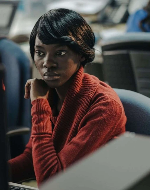 Portrait of an African woman with short, dark hair and a red sweater, looking slightly off-camera in a softly lit indoor office environment.