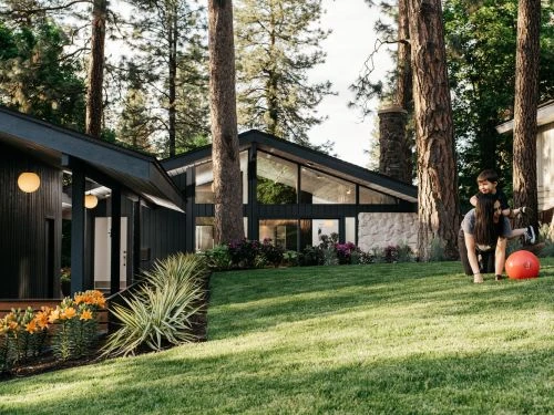 Adult female and child play on lawn in front of modern house with trees. Woman on hands and knees has child on her back.