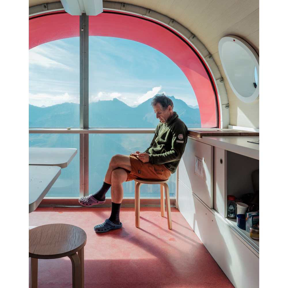 Man sitting on a stool inside a modern cabin, looking at a device with mountain range in the background through a large window.