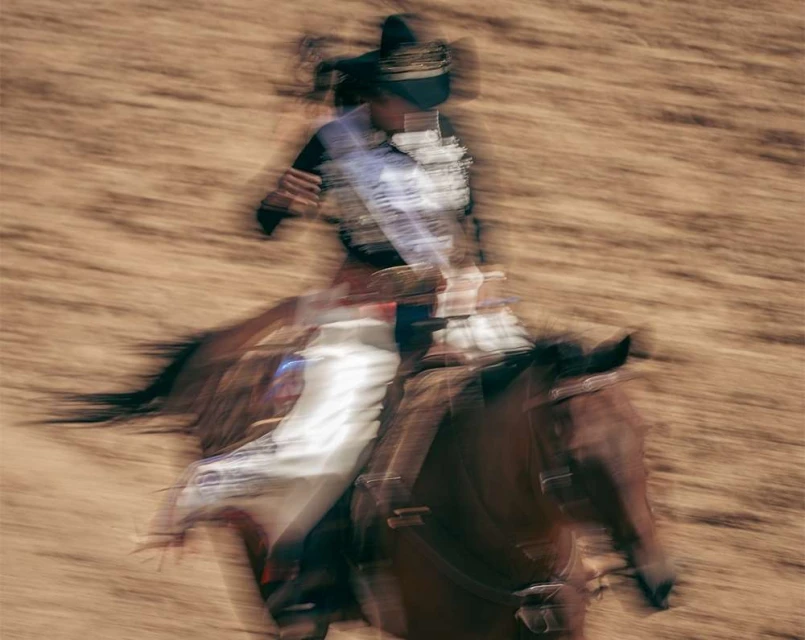 Motion blur photo of a horse and rider in ceremonial dress at speed on a dirt track with tan and brown colors dominating the scene.