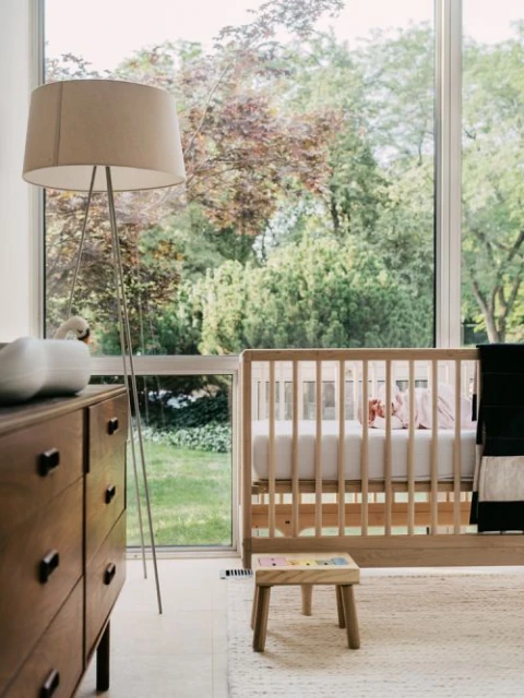 Nursery interior with a wooden crib, a wooden dresser with a modern lamp, a small wooden stool, and a large window overlooking greenery.