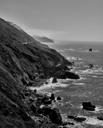 Monochrome landscape of a rugged coastline featuring cliffs, rocks, and ocean waves, captured in a black and white photograph.