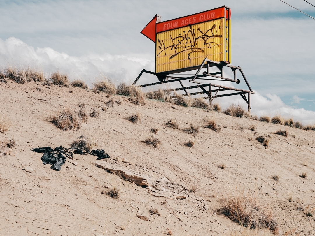 Vintage 'Four Aces Club' sign sits atop a sandy hill under a cloudy sky. The yellow sign with red trim features graffiti.