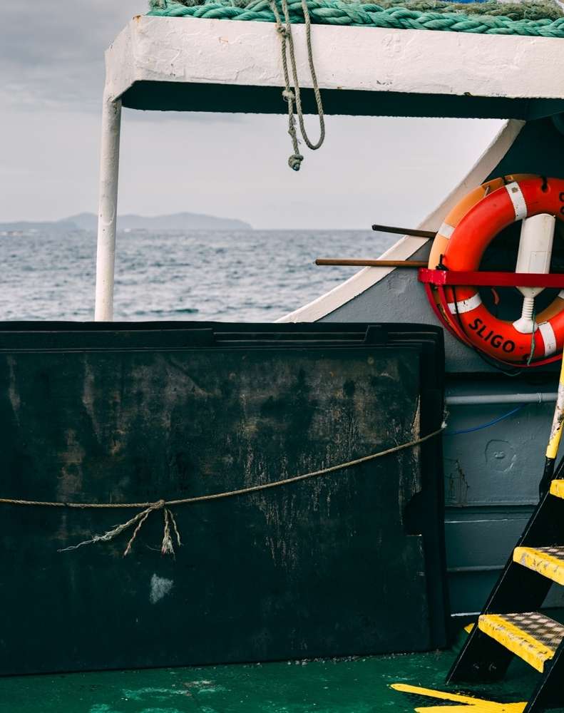 Marine scene featuring a black box on a ship deck, with a life preserver, sea, and distant land visible in the background.
