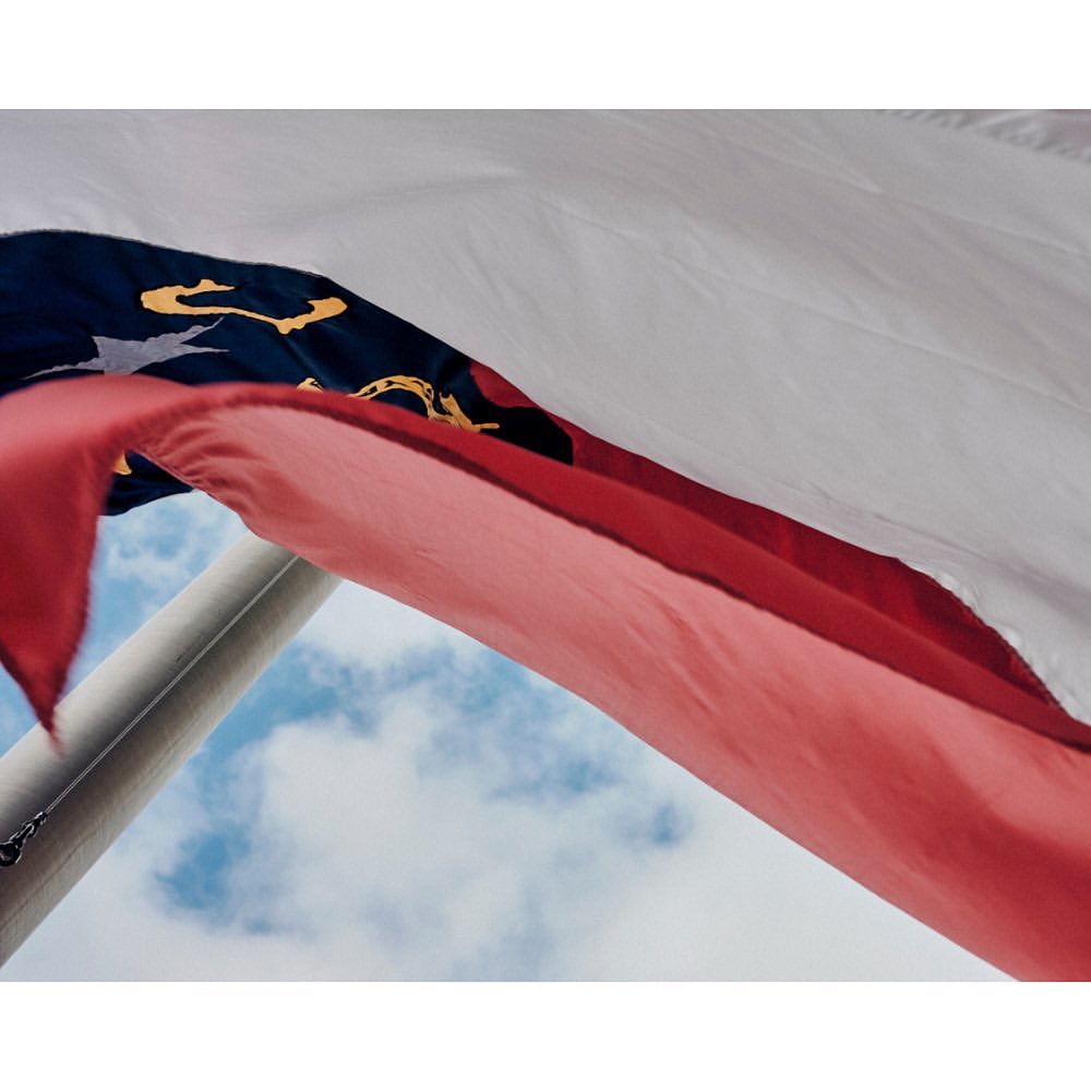 The North Carolina state flag waves against a bright, partially cloudy sky. The flag features a white stripe, a blue section, and a red stripe.