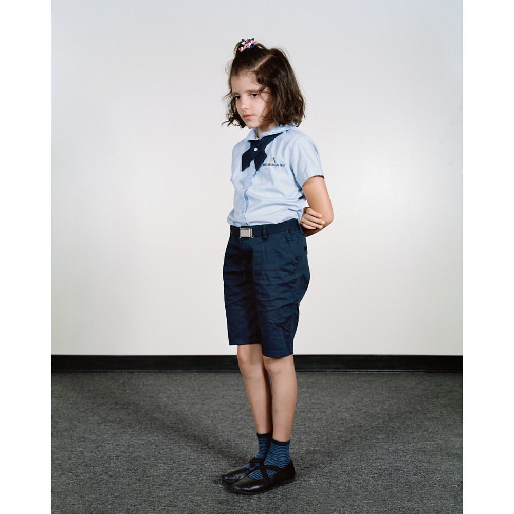 Young girl in a blue school uniform with a bow and dark shorts, standing indoors against a neutral background, portrait orientation.