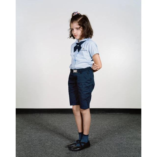 Young girl in a blue school uniform with a bow and dark shorts, standing indoors against a neutral background, portrait orientation.
