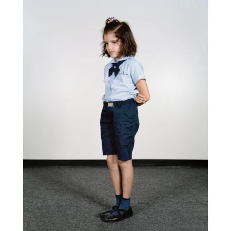 Young girl in a blue school uniform with a bow and dark shorts, standing indoors against a neutral background, portrait orientation.