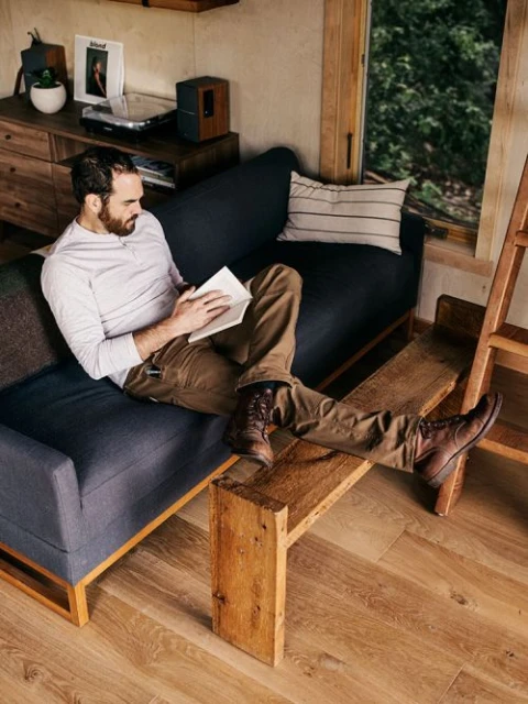 A man with a beard lounges indoors on a blue couch, reading a book in natural light, in a casual and relaxed setting, next to a window.