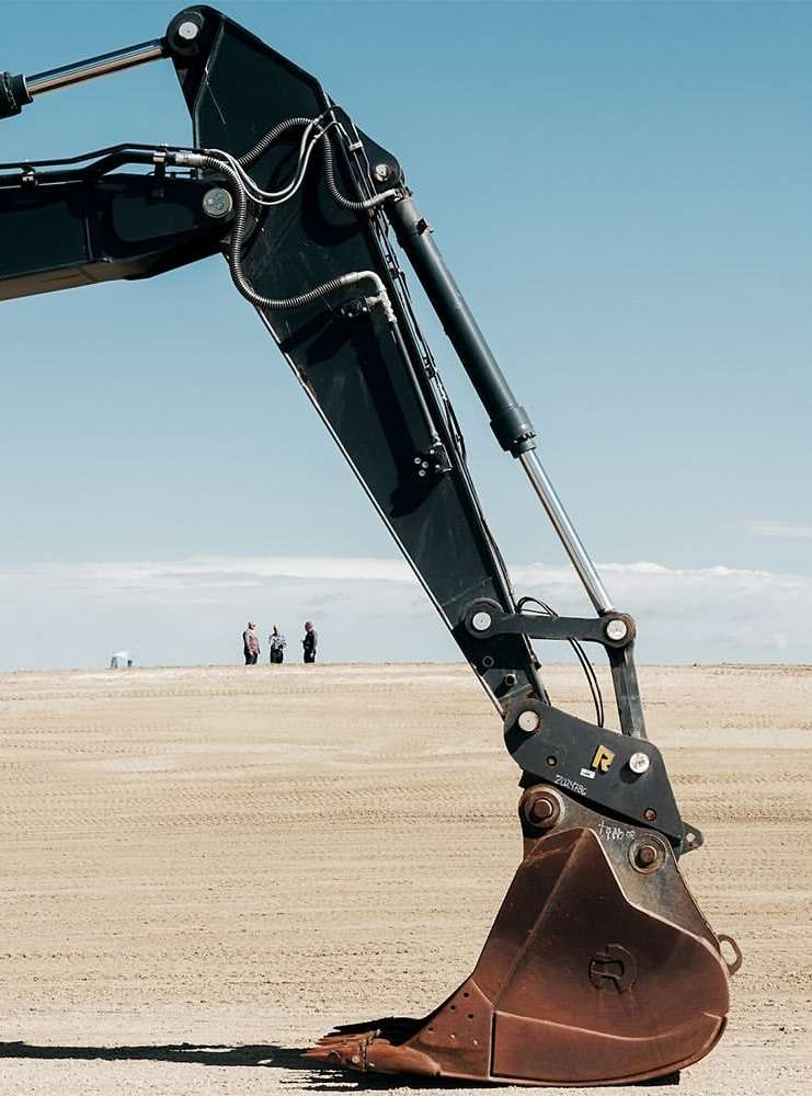 Excavator arm against a sandy landscape and a blue sky with three people standing in the distance, creating a stark contrast.