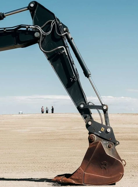 Excavator arm against a sandy landscape and a blue sky with three people standing in the distance, creating a stark contrast.