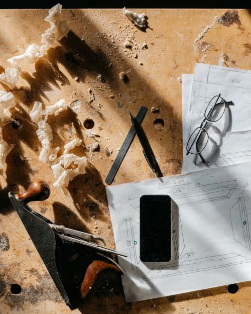 Overhead shot of a woodworking bench featuring a plane, smartphone, glasses, design plans, and wood shavings in natural light.