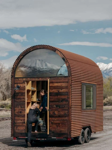 Two people enter a rustic tiny house on wheels with a corrugated metal exterior and a large arched window, set against a mountain backdrop.
