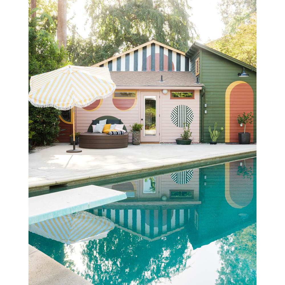 Outdoor poolside lounge area with a striped yellow umbrella, round daybed with pillows, modern painted buildings, and green backyard trees.