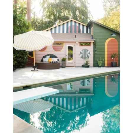 Outdoor poolside lounge area with a striped yellow umbrella, round daybed with pillows, modern painted buildings, and green backyard trees.