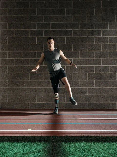Man with prosthetic leg balances on indoor track, demonstrating fitness and determination against a grey brick wall backdrop.