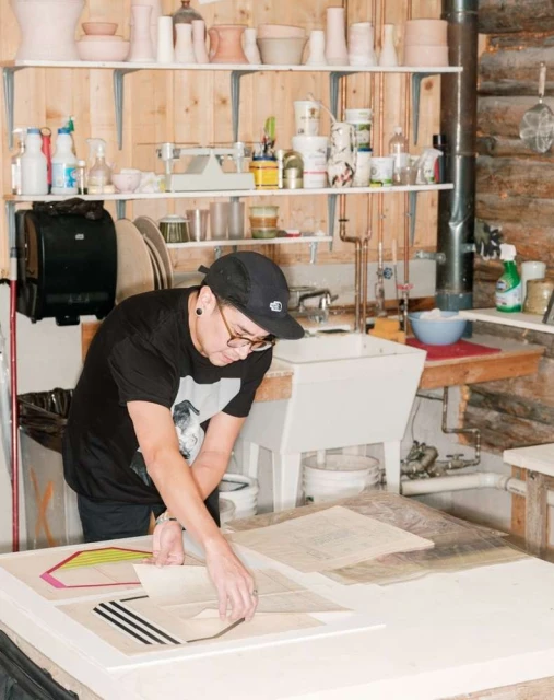 Adult working in a rustic studio, leaning over a table with artwork, surrounded by art supplies, pottery, and shelves.