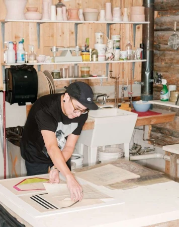Adult working in a rustic studio, leaning over a table with artwork, surrounded by art supplies, pottery, and shelves.