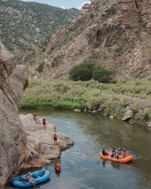 Whitewater rafting adventure on a sunny day, featuring a group of people in an orange raft navigating down a river in a rocky canyon.