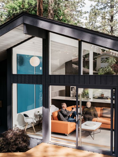 Two adult men relax in a modern living room visible through sliding glass doors, with a view of surrounding trees and an adjacent building.