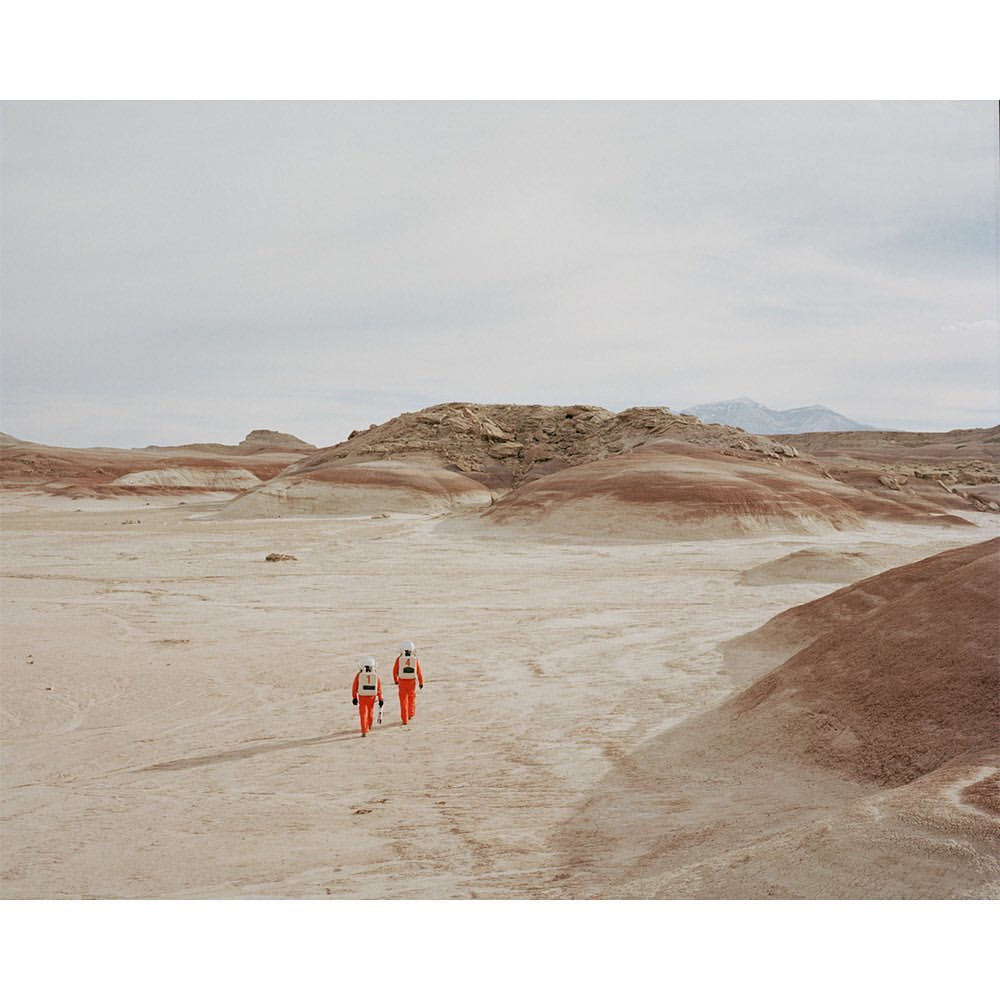 Two people in orange spacesuits walk across a desert landscape with red and tan rock formations under a cloudy sky, creating a sense of exploration.