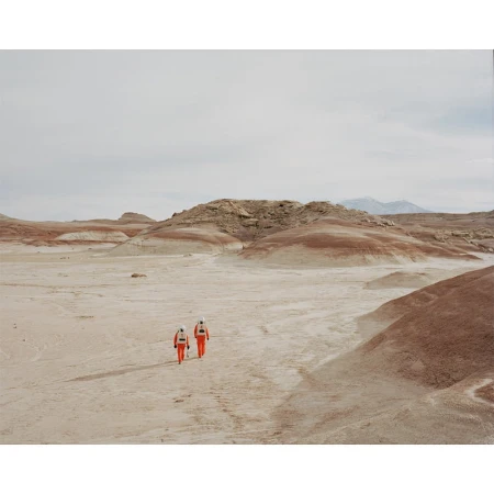 Two people in orange spacesuits walk across a desert landscape with red and tan rock formations under a cloudy sky, creating a sense of exploration.