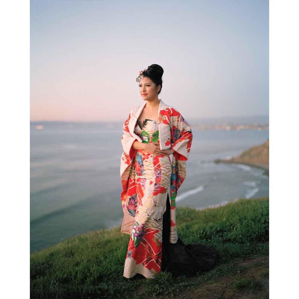 Woman in colorful kimono stands on a grassy hill overlooking the ocean at dusk, showcasing traditional Japanese fashion and natural beauty.