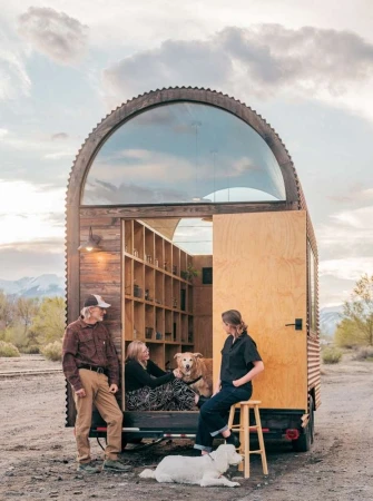Three people and two dogs pose outside of a unique tiny home on wheels in a rural setting with mountains and cloudy sky in the background.