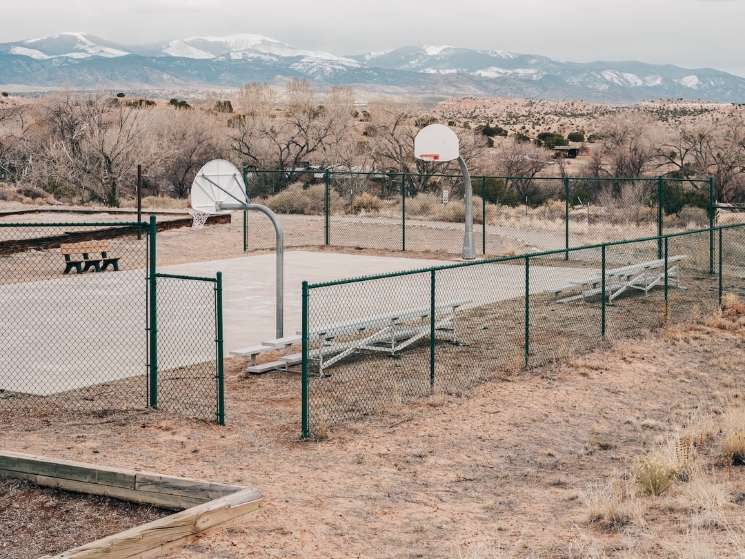 Outdoor basketball court with chain-link fence in desert setting, distant snow-capped mountains under cloudy sky, benches, and bare trees.