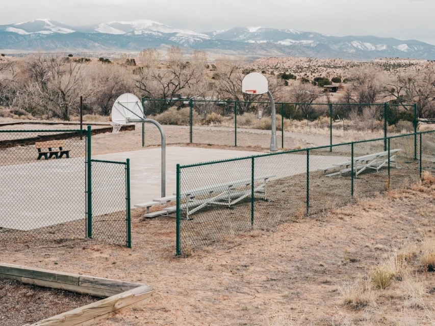 Outdoor basketball court with chain-link fence in desert setting, distant snow-capped mountains under cloudy sky, benches, and bare trees.